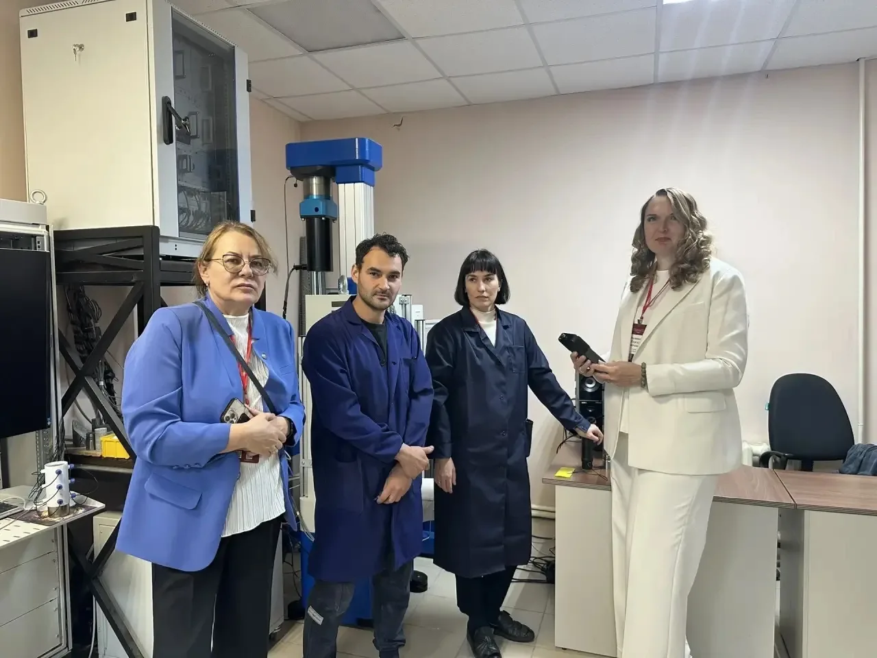 Четыре человека, трое в рабочих халатах, одна женщина в белом костюме, стоят в помещении с лабораторным оборудованием.