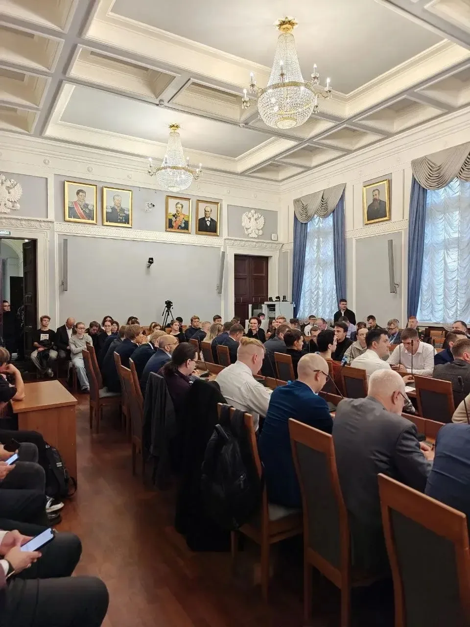 Зал, полный участников конференции, вид на президиум и портреты на стене.