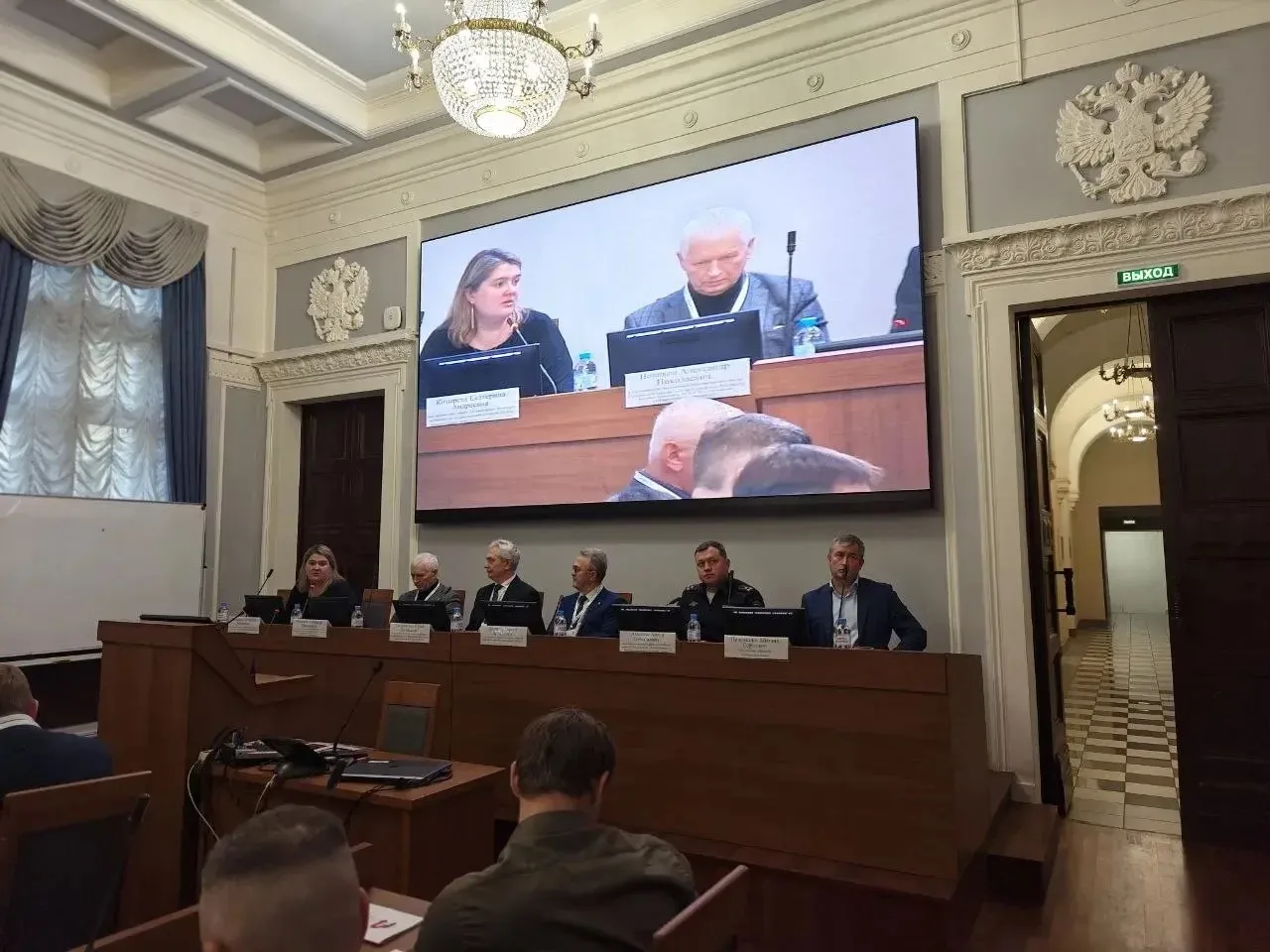 Участники конференции сидят за столами, на большом экране транслируются докладчики.