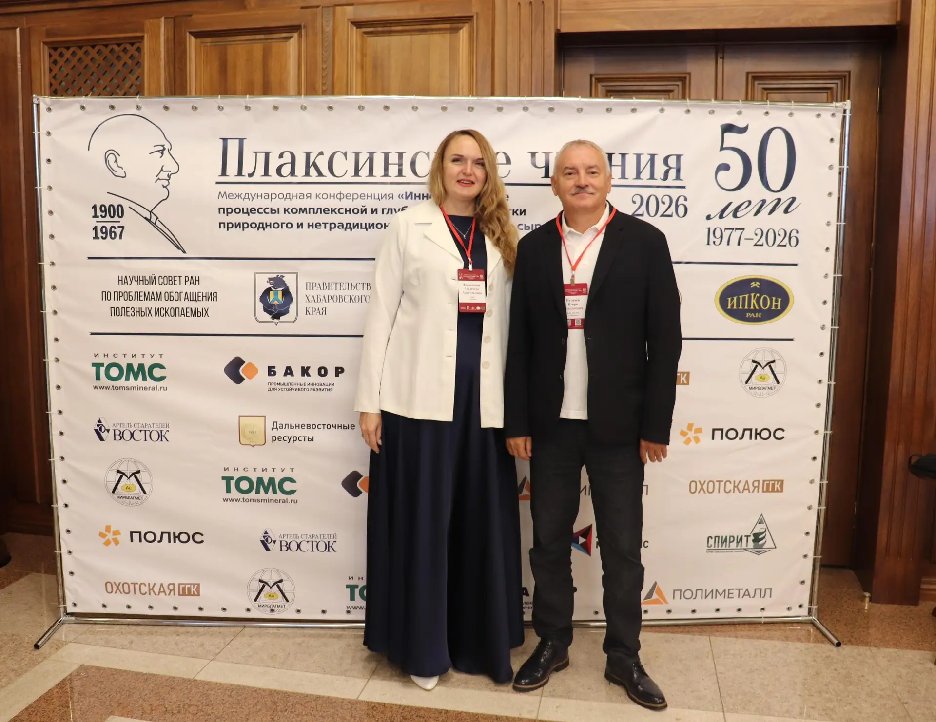 Two people stand in front of the 'Plaksin Readings' conference banner with logos of organisers and sponsors.