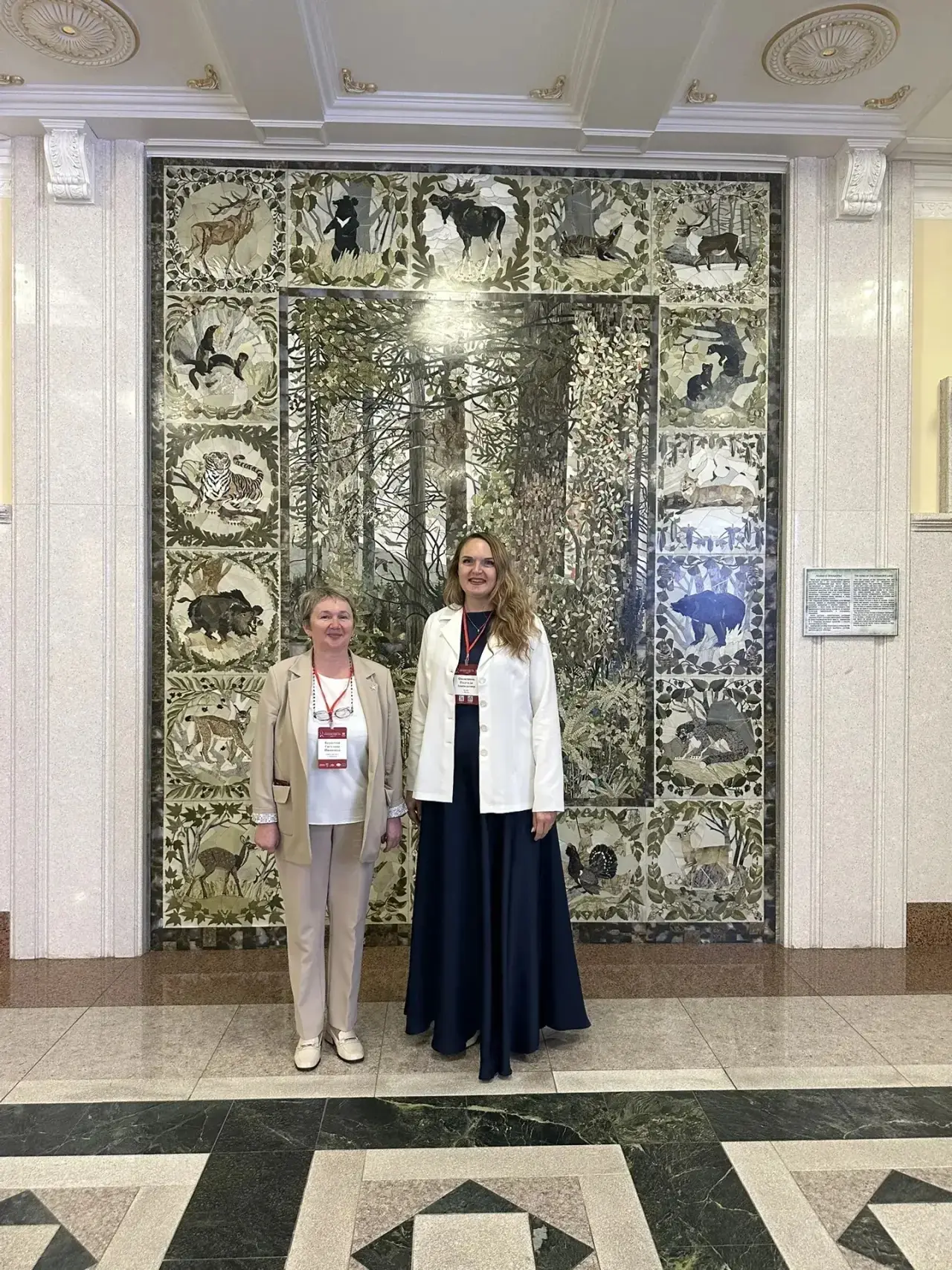 Two women stand indoors in front of a large mosaic panel depicting animals and a forest landscape.