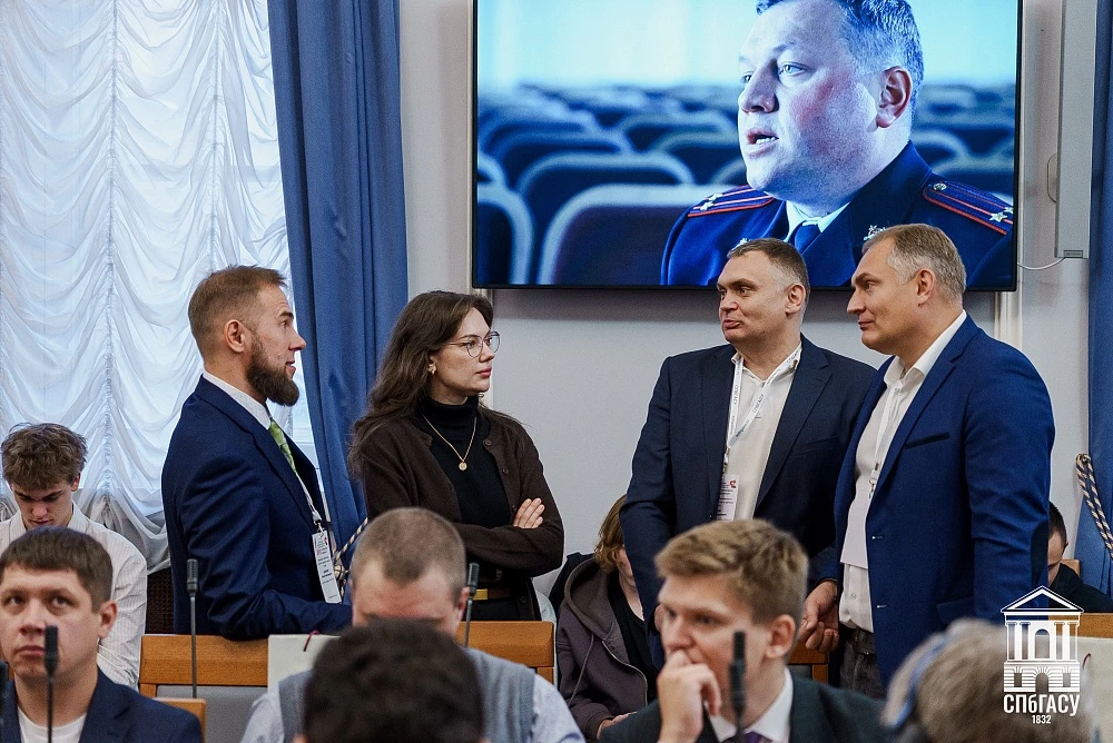 Группа людей обсуждает что-то в зале конференции, на экране транслируется крупный план докладчика.