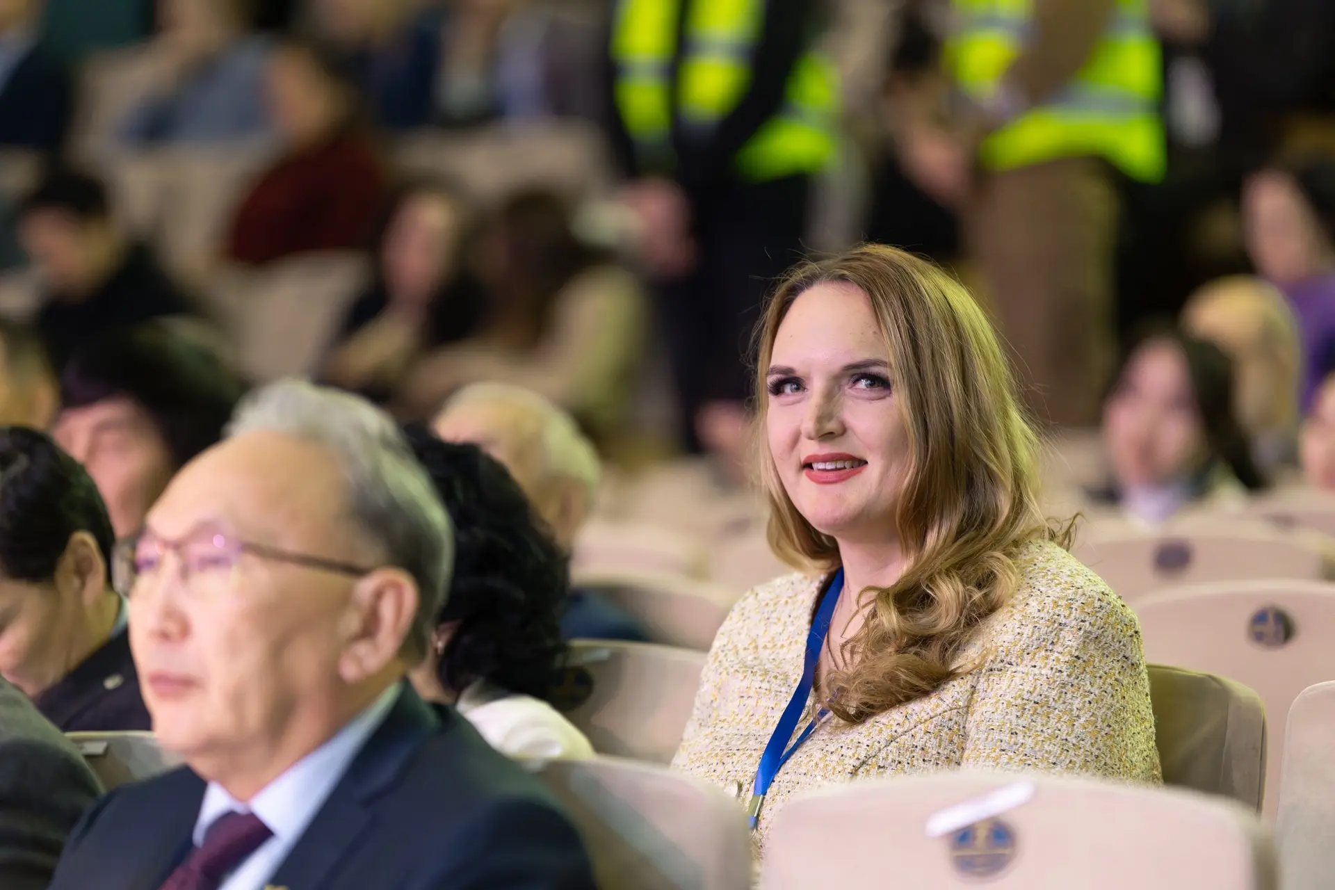 A woman with long blonde hair in a beige jacket sits at a conference.