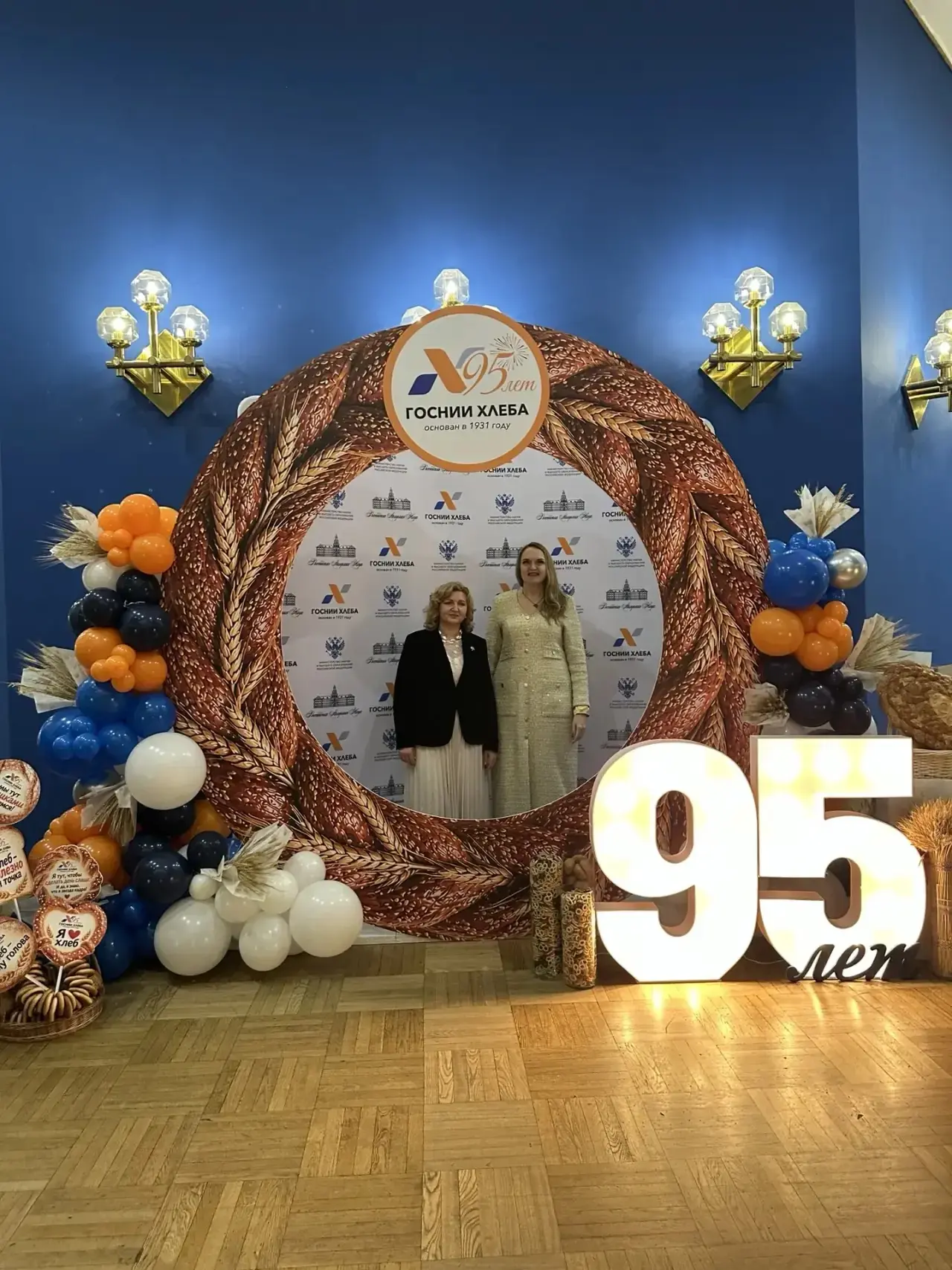 Two women stand in front of a stand with the inscription "95 years of GosNII Khleba" and decorations of wheat ears and balloons.