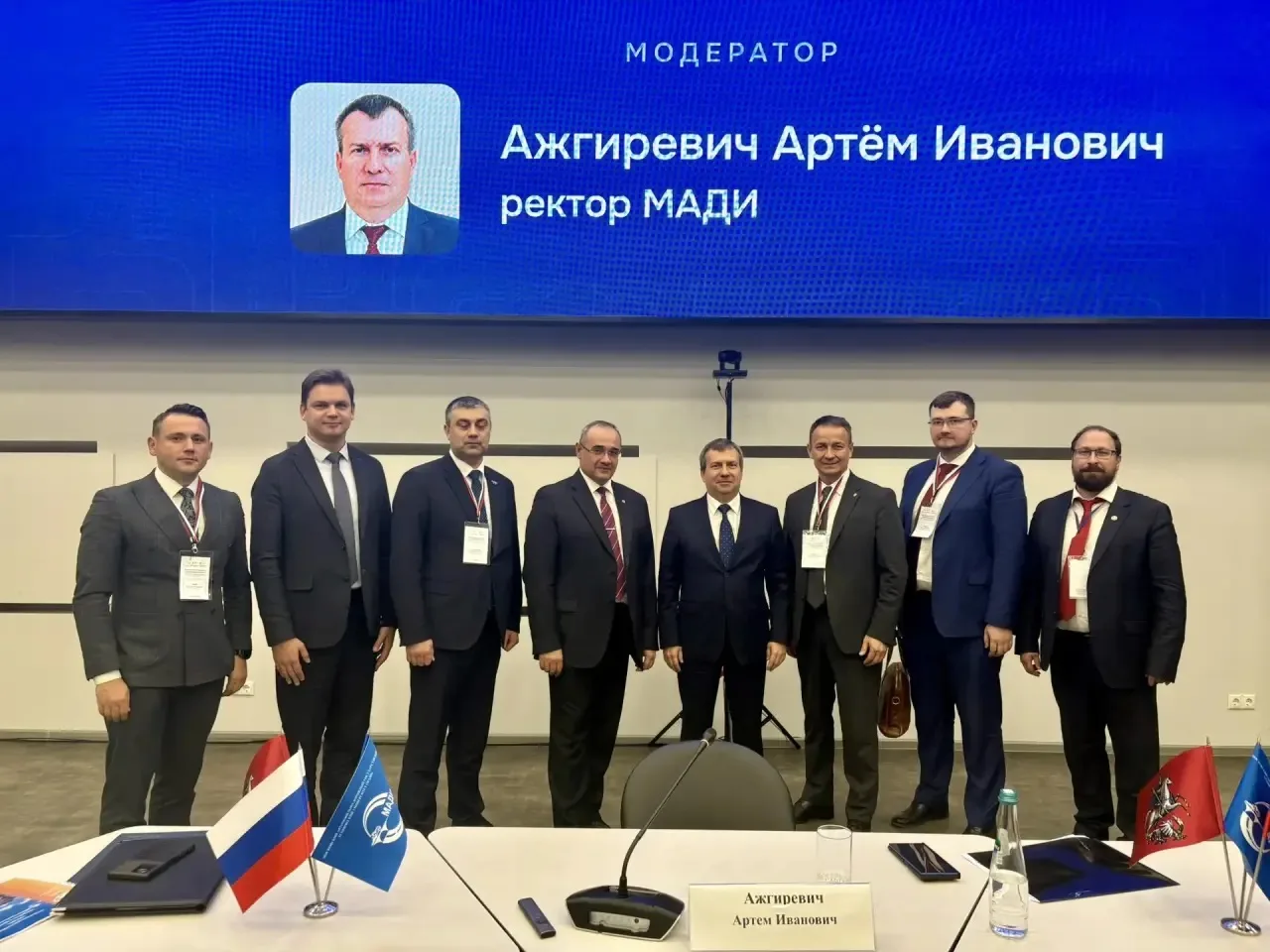 Группа мужчин в деловых костюмах, стоящих перед экраном с надписью 'МОДЕРАТОР Ажгиревич Артём Иванович ректор МАДИ'. На столе перед ними флаги России и МАДИ.