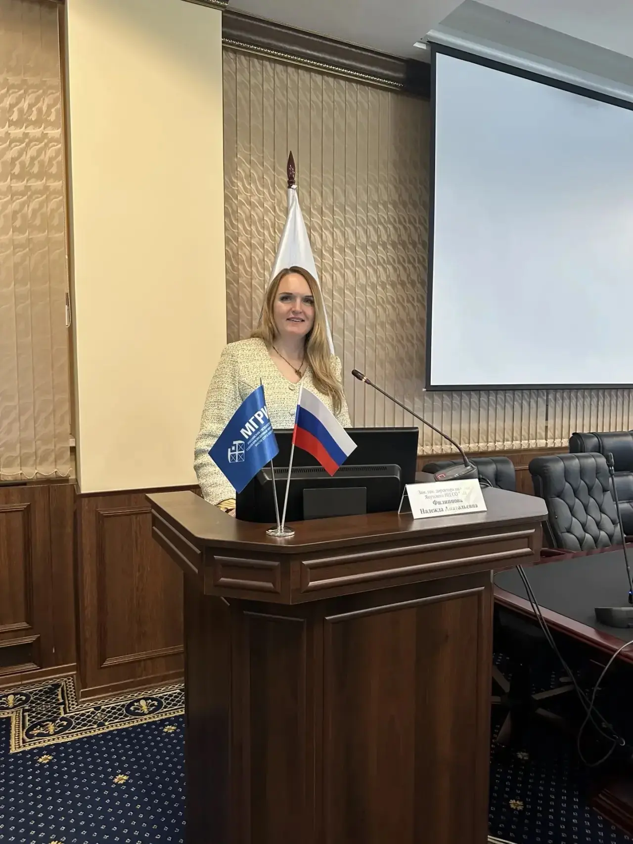 Nadezhda Filippova speaks at a podium with the flags of Russia and MGRI.
