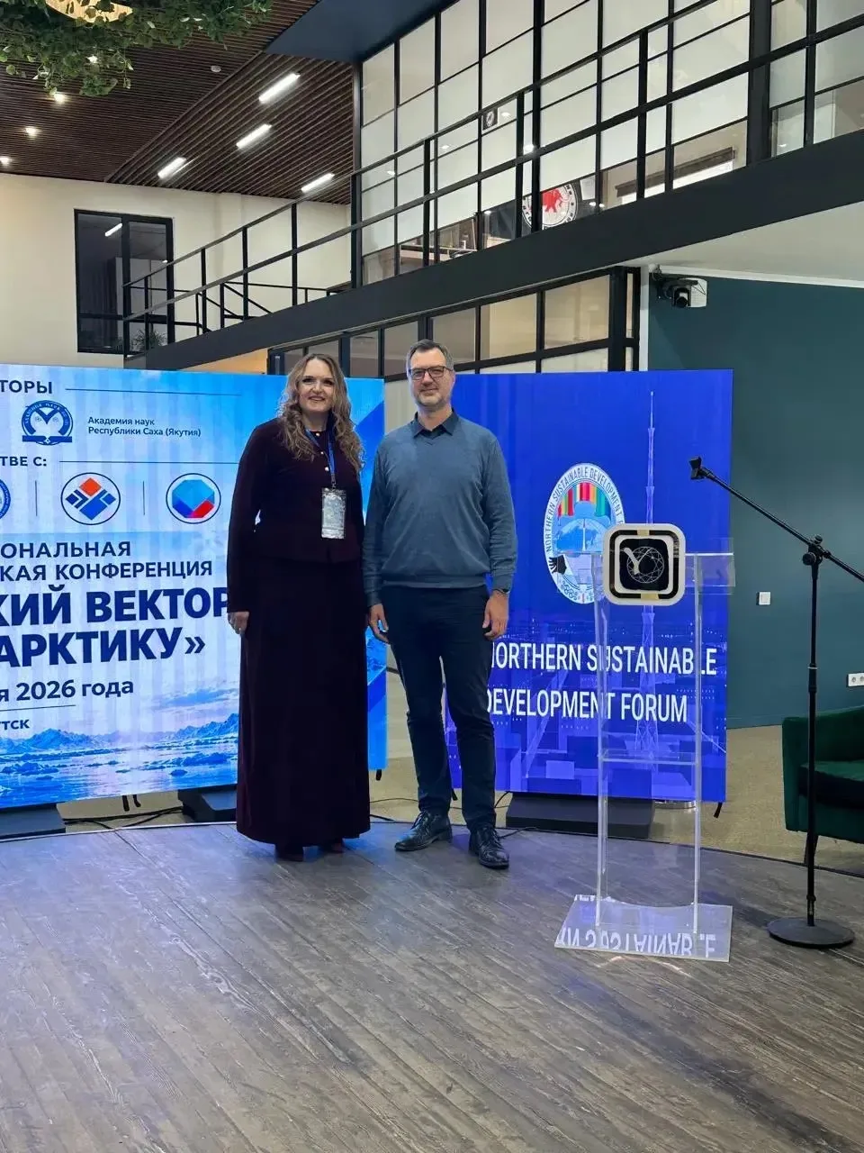 Надежда Филиппова и мужчина в сером свитере стоят на сцене на фоне баннера конференции.