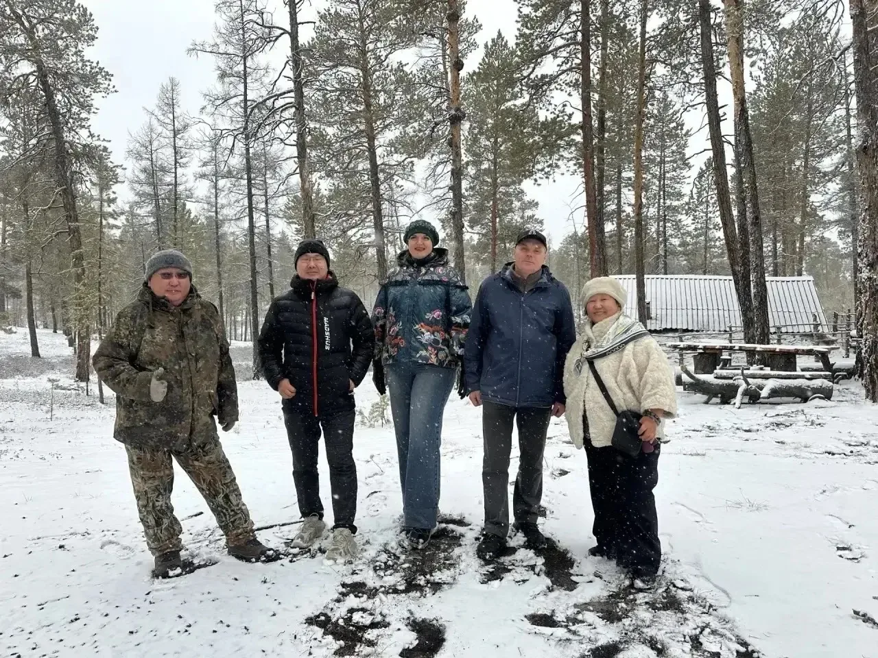 Группа из пяти человек стоит на заснеженной местности перед лесом.