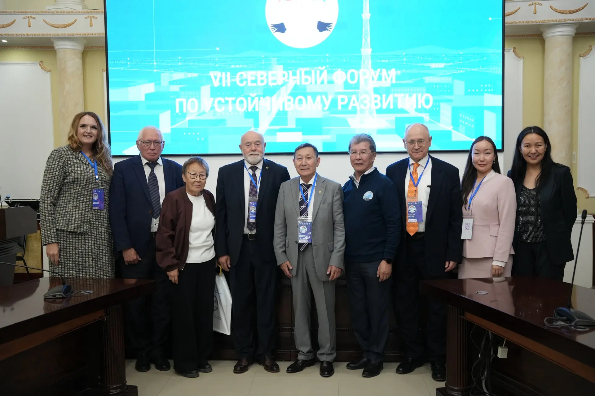 Nine people stand together in a room with a large screen in the background, showing the logo of the VII Northern Forum on Sustainable Development.